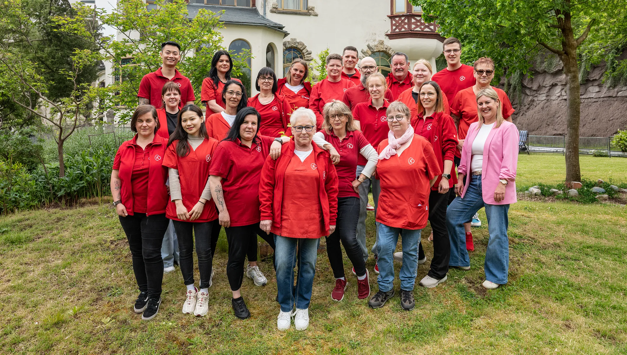 Team des Herzwohl Pflegedienstes in roten Dienstshirts bei einem Gruppenfoto im Grünen